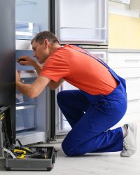 Male technician with screwdriver repairing refrigerator in kitchen